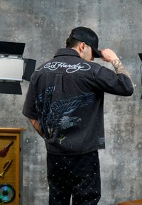 Homme portant une casquette noire et une veste noire avec un motif d'aigle et le texte « Ed Hardy », ajustant sa casquette à l'intérieur avec des lumières de studio et une décoration musicale à proximité.