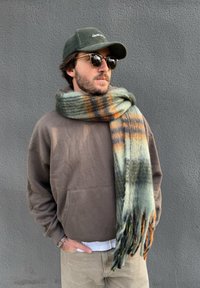 Écharpe à franges aux nuances de vert, orange et noir ; tissu doux et texturé. Sweat-shirt marron avec une poche kangourou, pantalon clair et casquette verte.