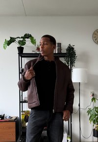 Veste marron zippée avec une finition texturée, portée sur un haut noir. Jeans foncés et accessoires complètent la tenue. Cadre intérieur avec des plantes.