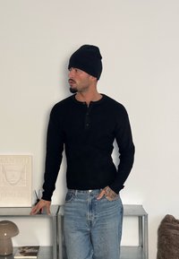 Chemise henley noire à manches longues avec une patte de boutonnage, portée avec un jean bleu clair ; le mannequin porte également un bonnet noir et présente de légers tatouages.