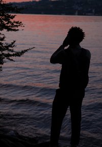 Henkilön siluetti seisoo veden äärellä, päällään tumma vaatetus ja reppu, auringonlasku heijastuu pinnasta.