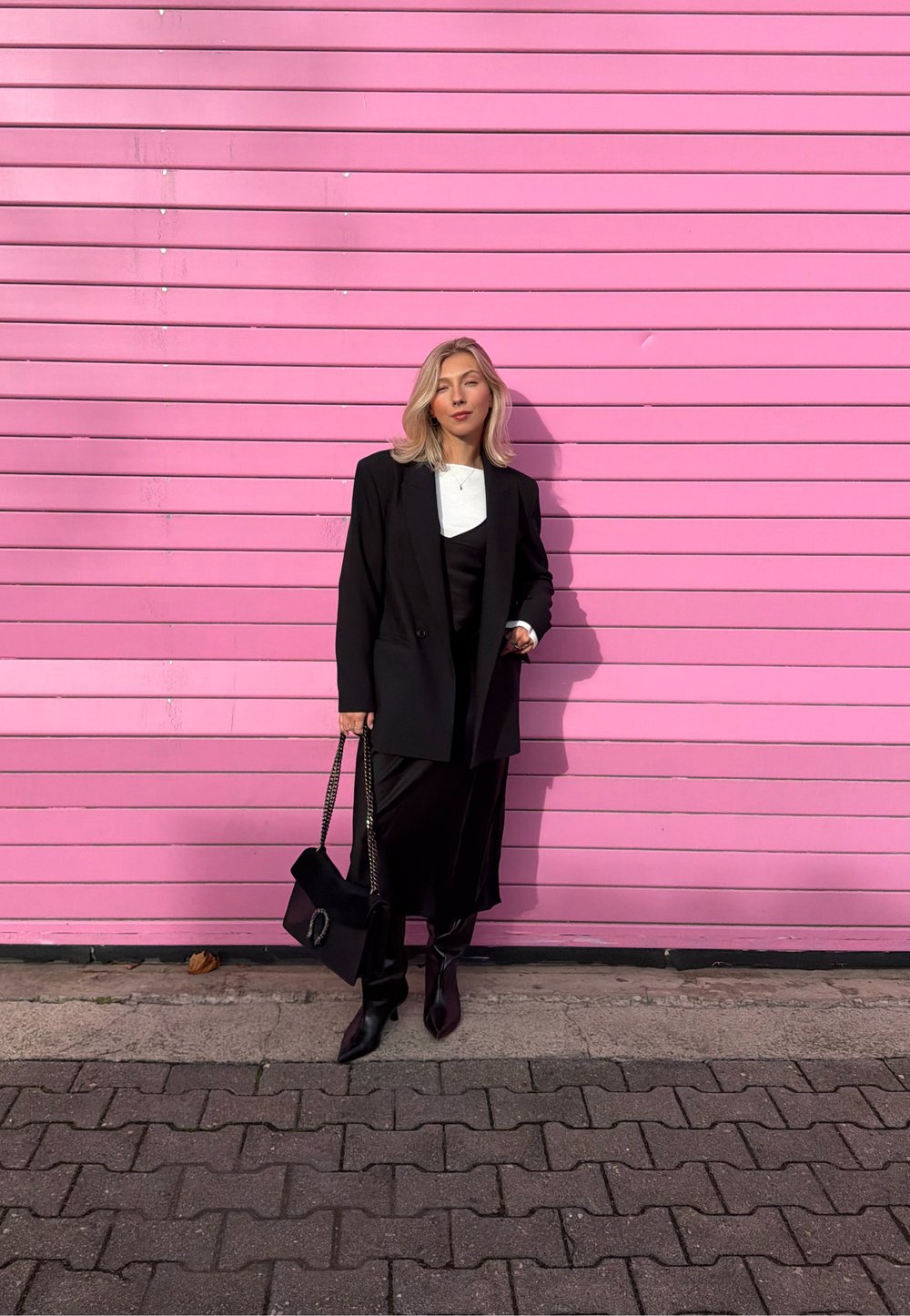 Blazer nero, top bianco, abito nero con spacco e stivali neri al ginocchio. Borsa nera con tracolla a catena. Sfondo con parete in doghe di legno rosa.