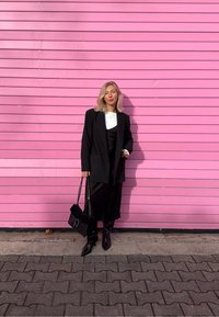 Blazer nero, top bianco, abito nero con spacco e stivali neri al ginocchio. Borsa nera con tracolla a catena. Sfondo con parete in doghe di legno rosa.