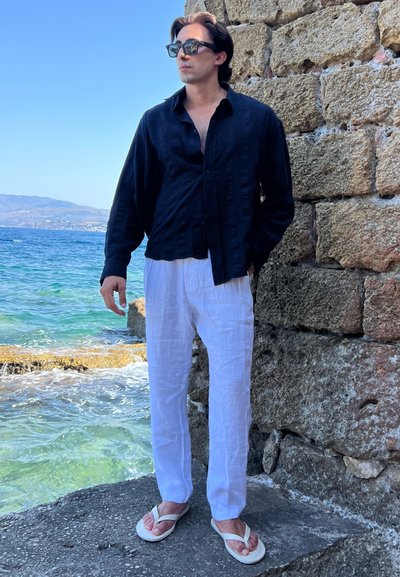 Camisa de lino negra de botones, pantalones de lino blancos y chanclas blancas, de pie cerca del agua con una pared de piedra al fondo.