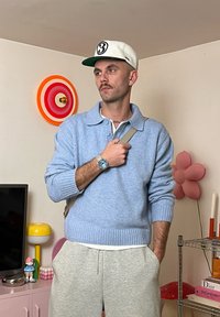 Svetlomodrý pletený sveter s rebrovanými manžetami, nosený cez biele tričko. Sivé tepláky a čiapka s číslom. Stojí v farebnej miestnosti.