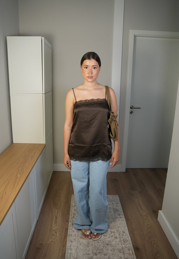 Jeune femme debout à l'intérieur portant un caraco en satin marron, un jean bleu clair, des sandales, et portant un sac à bandoulière marron.