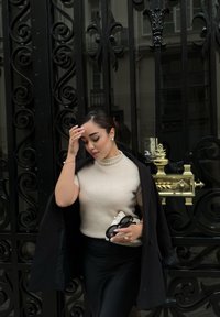 Maglione corto a maniche corte color crema, gonna nera a vita alta. Cappotto nero appoggiato sulla spalla. Collana e orecchini di perle. Tiene una piccola borsa.