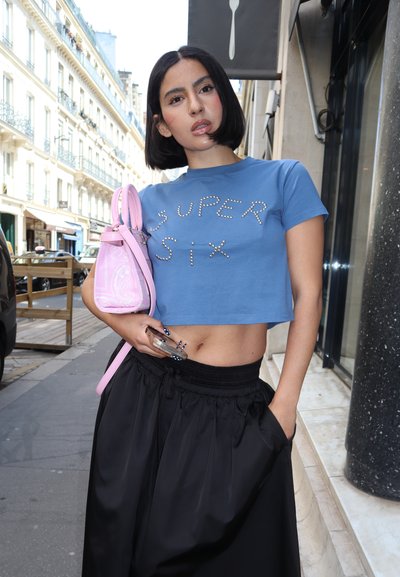 Camiseta azul cortada con "SUPER SIX" en cristales, combinada con una falda negra de cintura alta. Lleva un bolso rosa con un patrón paisley.
