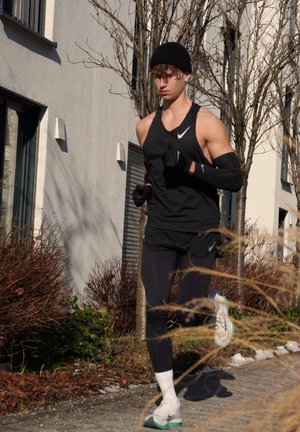 Joven vestido con ropa deportiva negra y un gorro, corriendo por una acera junto a árboles sin hojas y edificios residenciales bajo la luz del sol.