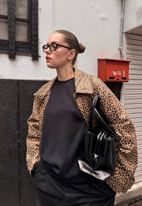 Leopárdmintás, bélelt dzseki széles gallérral, sötét texturált felsővel; fekete vödörtáskát tart a kezében. A háttérben egy piros postaláda látható.