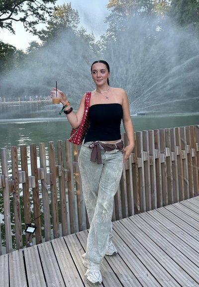 Top negro sin tirantes, pantalones de pierna ancha verde claro con un cinturón, bolso rojo con tachuelas, sosteniendo una bebida, con una fuente de agua de fondo.