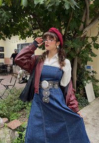 Denim dress with a fitted bodice and flared skirt, accented by a circular metal belt. Layered with a maroon bomber jacket and white blouse.