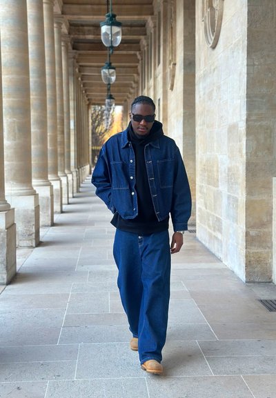 Hombre con chaqueta de mezclilla oscura, pantalones vaqueros, sudadera con capucha negra, gafas de sol y botas color camel caminando por un pasillo con columnas de piedra y faroles colgantes.