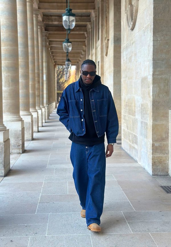 Mann iført mørk denimjakke, jeans, svart hettegenser, solbriller og beige støvler som går i en steinsøylekorridor med hengende lanterner.