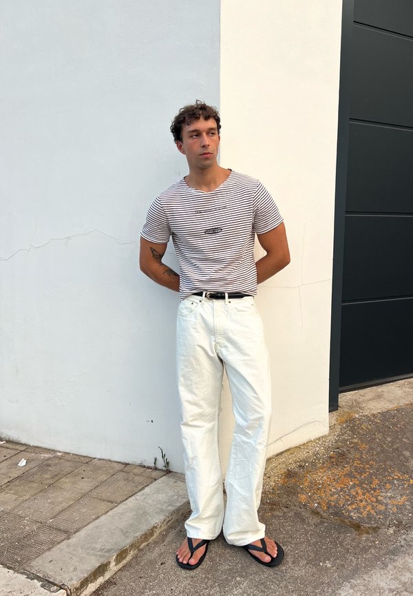 Mężczyzna o kręconych włosach, ubrany w pasiasty t-shirt, białe spodnie i czarne klapki, stoi oparty o ścianę z rękami za plecami.