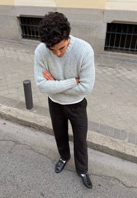 Maglione a coste grigio chiaro, pantaloni neri, scarpe di pelle nere con calzini bianchi. Uomo in piedi con le braccia incrociate in un contesto urbano.