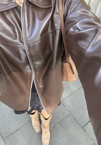 Giacca in pelle marrone con zip frontale, spalle strutturate e due tasche sul petto, abbinata a una gonna nera e stivali color cammello al ginocchio.