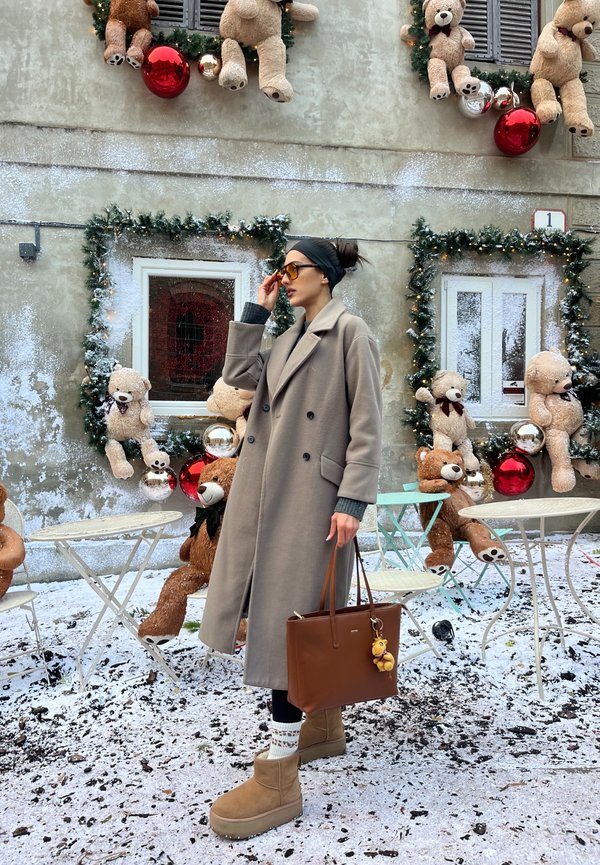 Femme en manteau long beige et bottes ajustant ses lunettes de soleil, debout près d'un mur enneigé décoré avec des ours en peluche et des ornements de Noël.