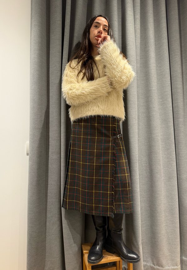 Femme debout sur un tabouret en bois portant un pull duveteux crème, une jupe à carreaux foncés et des bottes noires montantes, devant des rideaux gris.