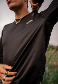 Persona che indossa una maglietta a maniche lunghe nera Adidas mentre tira il tessuto vicino alla vita, con anelli, collana e viso parzialmente visibile all'aperto.