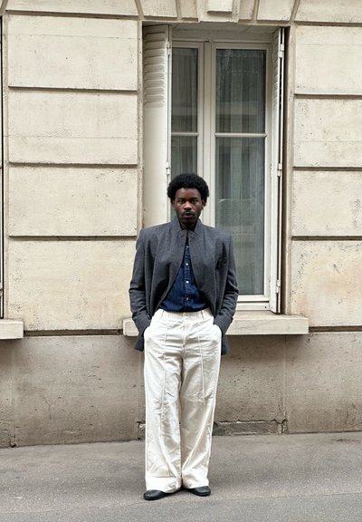 Hombre con las manos en los bolsillos, que lleva una blazer gris, camisa azul oscuro y pantalones blancos de pierna ancha, de pie frente a una ventana de un edificio beige.