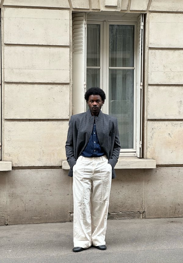 Άνδρας με τα χέρια στις τσέπες, φορώντας γκρι σακάκι, μπλε σκούρο πουκάμισο και λευκό παντελόνι με φαρδιά πόδια, στέκεται μπροστά από ένα μπεζ παράθυρο κτιρίου.