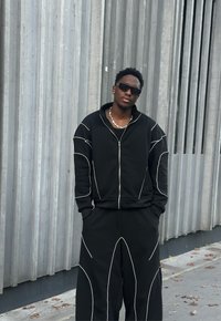 Sudadera negra con cremallera y costuras delineadas en blanco, combinada con pantalones de pierna ancha. Textura suave, ajuste relajado y adornos mínimos.