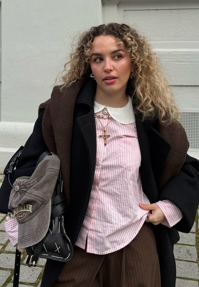 Joven con el cabello rizado, viste una camisa de rayas rosa, abrigo negro y lleva un suéter marrón sobre los hombros, sosteniendo una bolsa con una gorra enganchada.