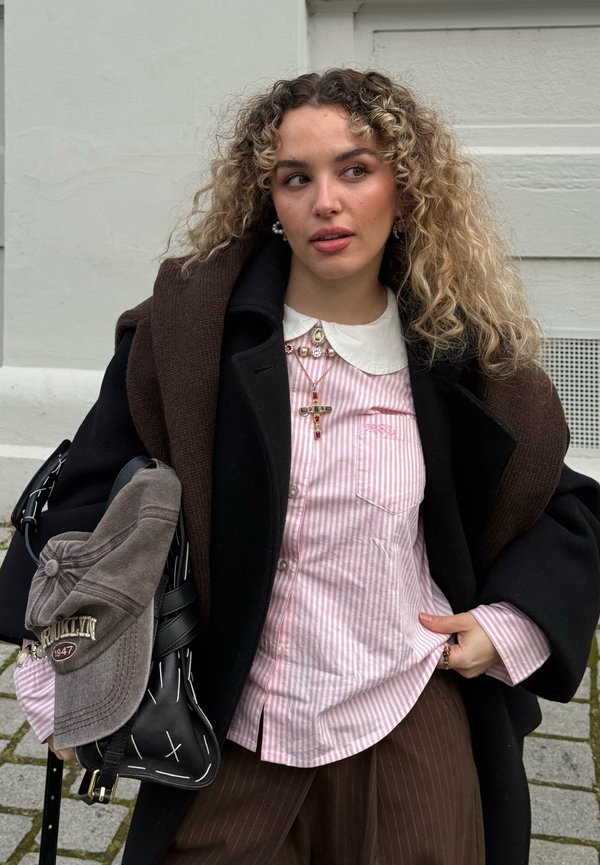 Ung kvinde med krøllet hår iført en stribet lyserød skjorte, sort frakke, brun sweater lagt over skuldrene, holder en taske med en kasket fastgjort.