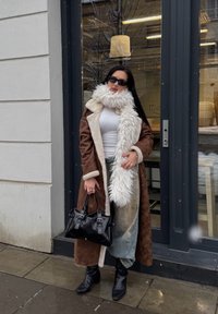 Ilgas rudas paltas su baltu kailio pamušalu, balta palaidinė, išblukusios mėlynos džinsinės kelnės, juoda rankinė su sidabro detalėmis ir juodi aulinukai.