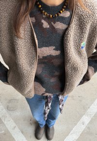 Brun fleecejakke med en camouflage-mønstret sweater under, blå jeans og brune ankelstøvler. Tilbehør med en perlekæde.