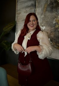 Burgundia värvi korduroidkleit valge pluusiga, millel on lips. Hoides väikest, ümarat burgundia värvi käekotti. Taustal on taim ja kunst.
