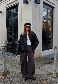 Chaqueta de cuero negro extragrande, camiseta blanca, pantalones anchos marrones, boina negra y gafas de sol, de pie sobre adoquines cerca de una cafetería.