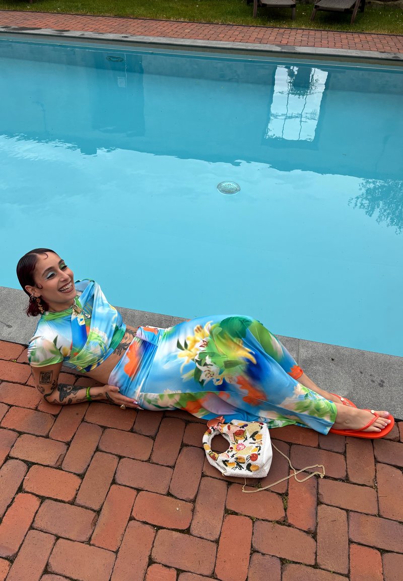 Personne portant une tenue colorée à deux pièces à fleurs et des sandales orange se détendant sur le bord de la piscine en briques près d'un sac brodé blanc.