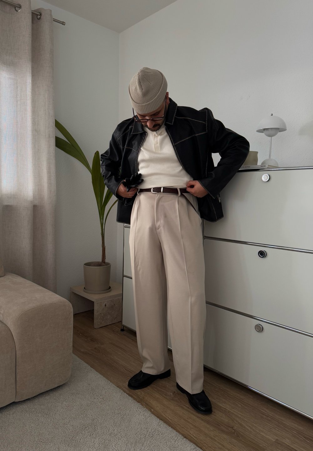 Chaqueta de cuero negra con costuras blancas, suéter de canalé crema, pantalones beige claros y zapatos negros, en un interior minimalista.