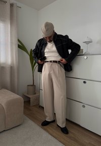 Chaqueta de cuero negra con costuras blancas, suéter de canalé crema, pantalones beige claros y zapatos negros, en un interior minimalista.
