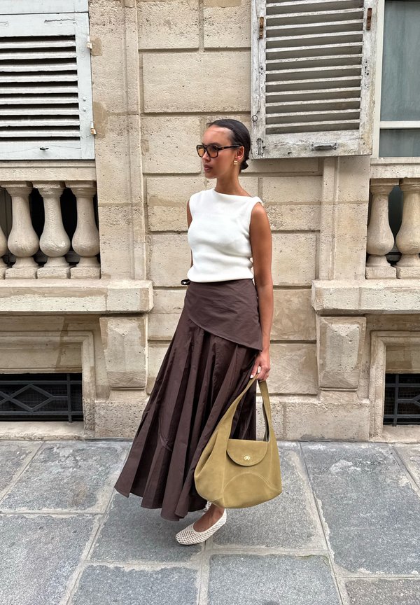 Femme en haut blanc sans manches et longue jupe marron tenant un sac à main olive, debout près d'un bâtiment en pierre avec des fenêtres à volets.