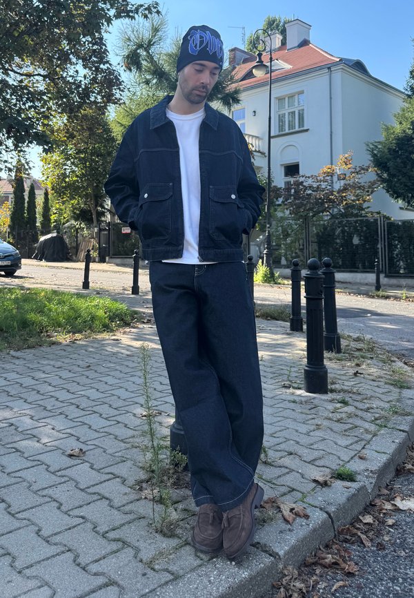Hombre que lleva una chaqueta negra, vaqueros anchos y un gorro negro, de pie en una acera cerca de una calle con casas y árboles al fondo.