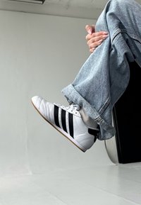 Witte sportschoen met zwarte accenten, met een gestructureerde bovenkant en drie parallelle strepen. Draagt bij lichte blauwe denim, omgeslagen broeken.