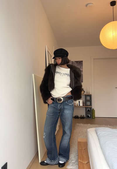 Mujer con chaqueta de piel negra, camiseta blanca con diseño gráfico, vaqueros azules con una hebilla grande en el cinturón, sombrero negro y zapatos negros, de pie en un dormitorio.