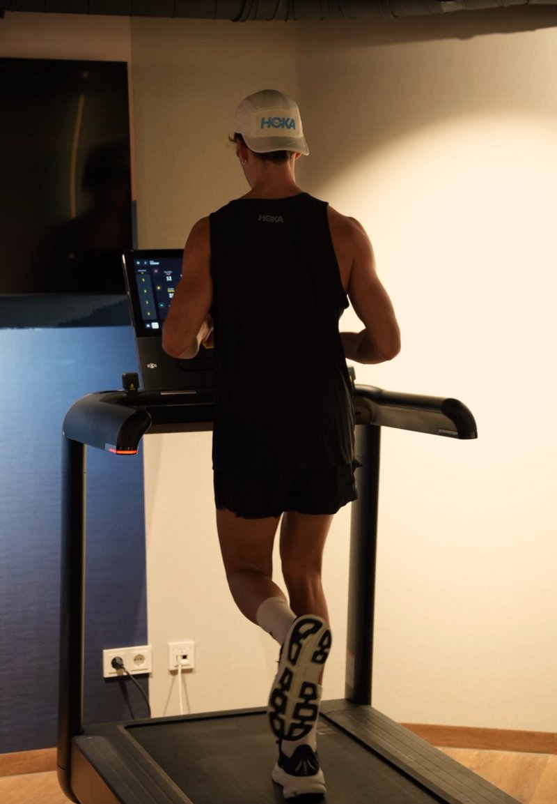 Tapis roulant noir avec console numérique, utilisateur portant un débardeur noir et un short, chaussettes blanches, en train de courir, semelle en caoutchouc texturée visible.