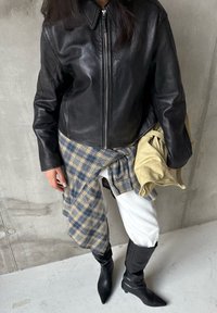 Veste en cuir noire à col montant, portée sur une chemise à carreaux bleue et jaune, associée à un pantalon blanc et des bottes noires à hauteur des genoux.