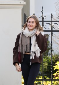 Καφέ oversized σακάκι, μαύρο crop top και ψηλόμεσα κολάν. Μπεζ και γκρι κασκόλ με φούντες. Πλεξιγκέιτ τσάντα σε μπορντό χρώμα. Πράσινη φυλλωσιά στο φόντο.