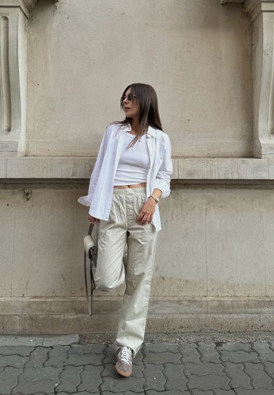 Camisa blanca de botones sobre un top corto blanco, combinada con pantalones texturizados beige claros y zapatillas grises. Sosteniendo un bolso pequeño.
