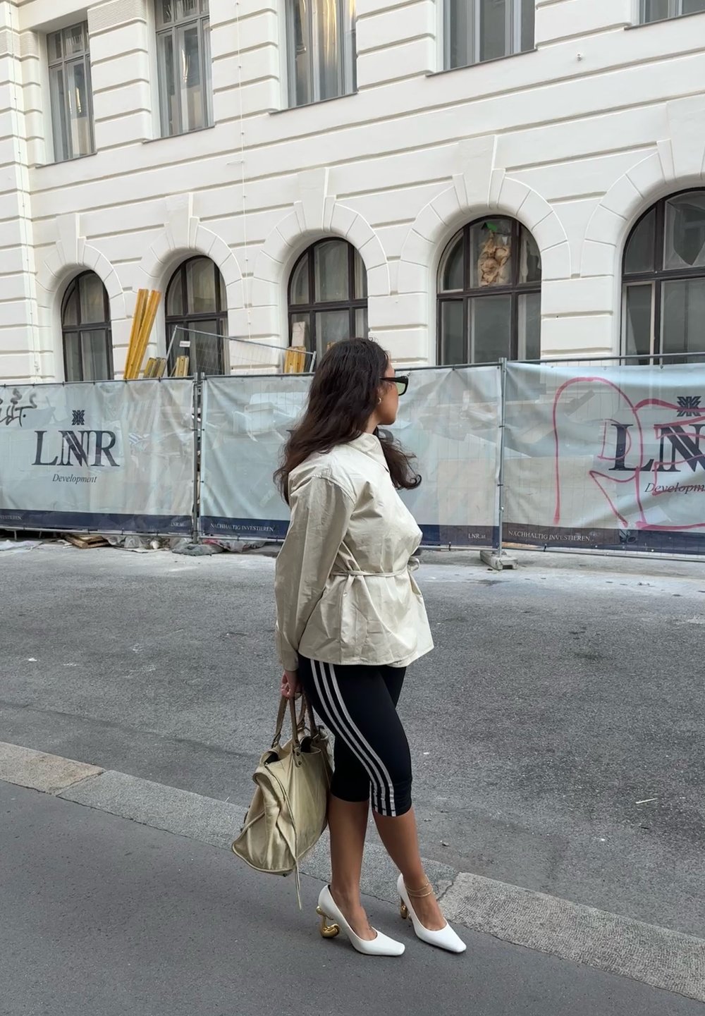 Kvinde i beige jakke, sorte leggings med hvide striber og hvide højhælede sko, holder en beige taske, står på fortovet nær en byggepladsbarriere.