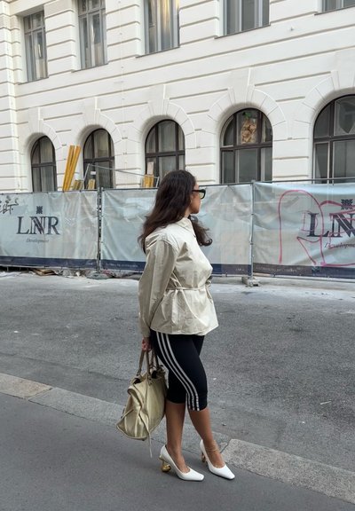 Mujer con chaqueta beige, leggings negros con rayas blancas y tacones blancos sosteniendo un bolso beige, de pie en la acera cerca de una barrera de construcción.