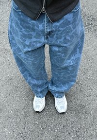 Laisvai krentančios mėlynos džinsinės kelnės su subtiliu kamufliažo raštu, derinamos su baltais sportiniais bateliais ant pilko šaligatvio. Tekstūra atrodo dėvėta, bet nepažeista.