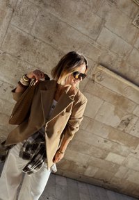 Femme portant un blazer beige oversize, des lunettes de soleil, un pantalon blanc et une chemise à carreaux nouée à la taille, tenant un sac tissé devant un mur en pierre.