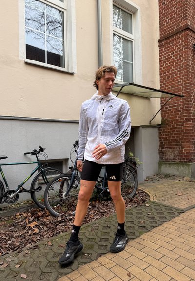 Chaqueta blanca y ligera con rayas negras, combinada con pantalones cortos de ciclismo negros. Zapatillas deportivas negras, caminando sobre un camino de piedra con patrón.