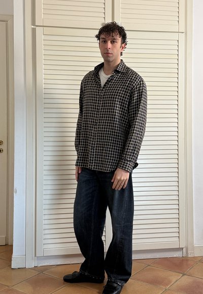 Joven de pie en el interior, vistiendo una camisa de cuadros en blanco y negro, jeans anchos oscuros y zapatos negros.